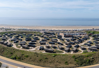 Vista aérea de casas modernas junto a la playa con vehículos aparcados y un mar tranquilo al fondo.
