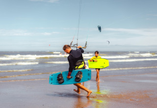 Dos personas con tablas de kitesurf caminan por la playa junto a Sea Lodges Zandvoort 3 en los Países Bajos.