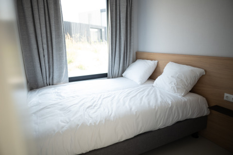 Dormitorio luminoso con cama individual, ropa de cama blanca, dos almohadas y gran ventana en Sea Lodges Zandvoort 1.