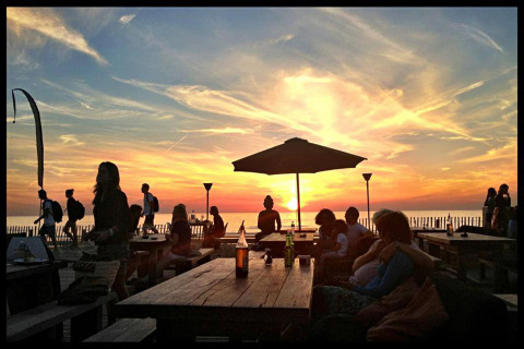 Zonsondergang bij glampingverblijf, mensen relaxen aan picknicktafels en genieten van het zeezicht.