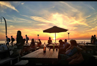 Zonsondergang bij glampingverblijf, mensen relaxen aan picknicktafels en genieten van het zeezicht.