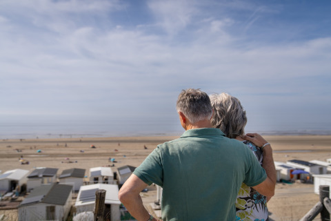Una pareja mayor contempla el mar y la playa desde una casa de playa bajo un cielo azul despejado.