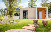 Lodge moderno con sedute in tronchi di legno in un giardino verde, circondato da alberi e luce naturale.