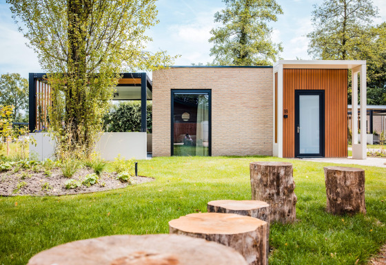 Moderne lodge med træstammer som siddepladser i en grøn have omgivet af træer og naturligt lys.