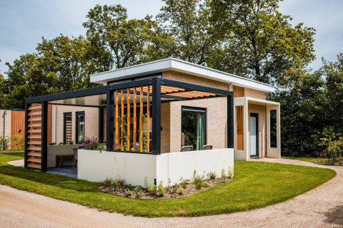 Lodge moderno con ampia terrazza, pergolato e giardino, circondato da alberi e prato verde.