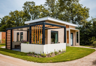 Lodge moderno con ampia terrazza, pergolato e giardino, circondato da alberi e prato verde.