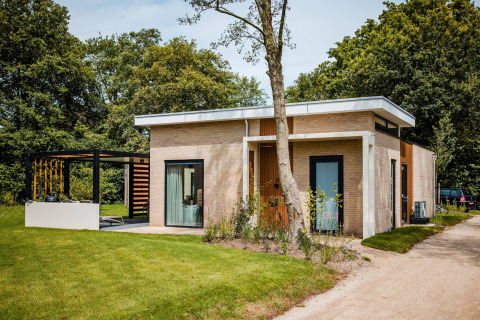 Lodge moderno con grandi finestre, tetto piano e patio, immerso in un giardino verde tra gli alberi.