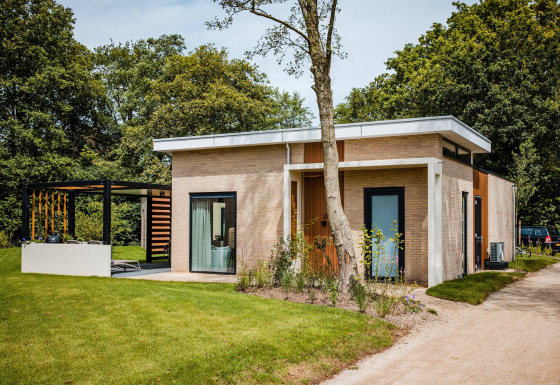 Moderne lodge med fladt tag, store vinduer og terrasse, omgivet af grøn have og træer i naturen.
