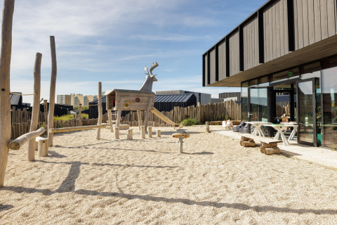 Parque infantil al aire libre con figuras de madera junto a una casa de playa moderna y edificios al fondo.