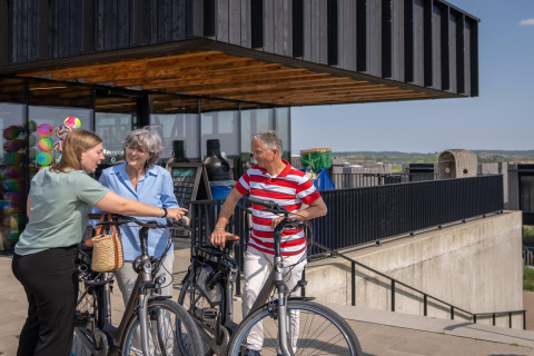 Tres adultos conversan afuera de una casa de playa moderna con bicicletas, disfrutando un día soleado.