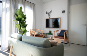 Helles Wohnzimmer mit grĂĽner Couch, Sessel, Wand-TV und groĂźer Pflanze in Sea Lodges Zandvoort 9, Niederlande.