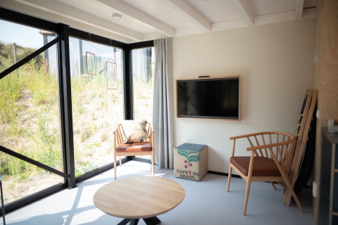 Lichte leefruimte in Sea Lodges Zandvoort 9 met houten stoelen, groot raam en zicht op de duinen, Nederland.