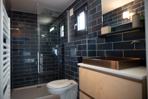 Moderne badkamer met donkerblauwe tegels, glazen douche, toilet en raam in Sea Lodges Zandvoort 6, Nederland.