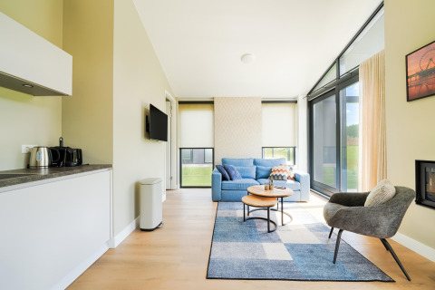 Salon lumineux avec canapé bleu, fauteuil, grandes fenêtres et kitchenette dans un gîte près des lacs Kaag.