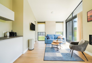 Salon lumineux avec canapé bleu, fauteuil, grandes fenêtres et kitchenette dans un gîte près des lacs Kaag.