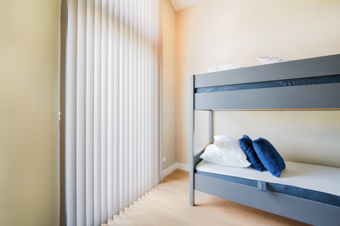 Chambre moderne avec lits superposés gris, coussins blancs et bleus, et grands stores verticaux aux fenêtres.