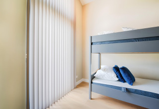 Chambre moderne avec lits superposés gris, coussins blancs et bleus, et grands stores verticaux aux fenêtres.