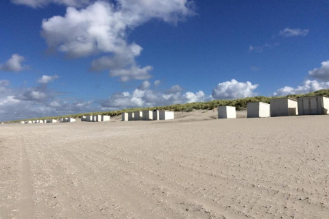 White glamping cabins lined up on a sandy beach with dunes and a blue sky with scattered clouds.