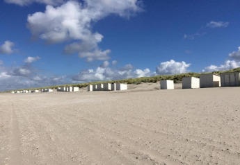 Bungalow glamping bianchi su una spiaggia sabbiosa vicino a dune, con cielo blu e nuvole sparse.