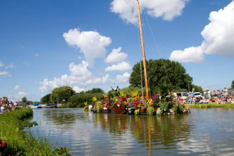A flower-decorated raft floats past spectators on a sunny day at a canal near a glamping site.