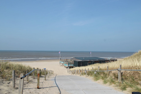 Een pad tussen duinen leidt naar strandpaviljoen De Schone Leij aan een zonnig Nederlands strand.
