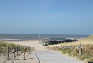 Een pad tussen duinen leidt naar strandpaviljoen De Schone Leij aan een zonnig Nederlands strand.