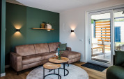 Moderne lodge-stue med brun sofa, grøn væg, runde borde og udgang til terrasse med træafskærmning.