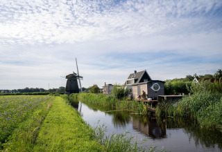 Kleines Haus am Kanal mit Windmühle im Hintergrund in den Niederlanden bei Küstenlandschaft.