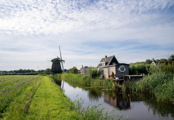 Tiny house aan een kanaal met windmolen op de achtergrond nabij de kust in Nederland, zonnige dag.