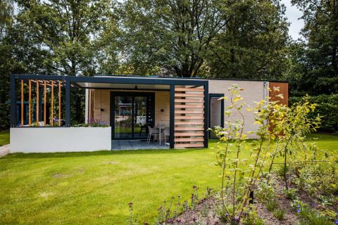 Modern lodge met ruime veranda en tuin, te midden van bloemen, gras en bomen in een natuurlijke sfeer.