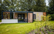 Moderne lodge med terrasse, grøn græsplæne og blomsterbede, omgivet af træer i naturligt miljø.