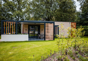 Moderne lodge med terrasse, grøn græsplæne og blomsterbede, omgivet af træer i naturligt miljø.