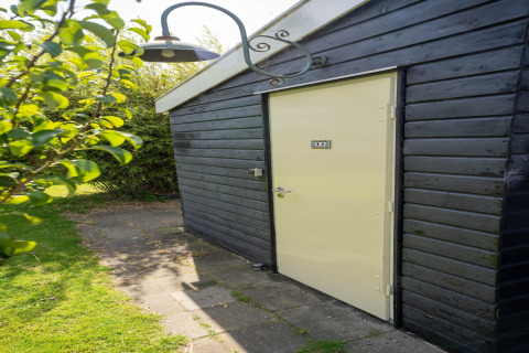 Eingangstür eines kleinen Hauses in den Niederlanden, schwarze Holzverkleidung und Garten im Sonnenlicht.