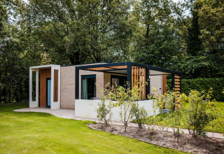 Moderne Lodge mit Terrasse im Grünen, umgeben von Bäumen und gepflegtem Rasen im Sommerlicht.