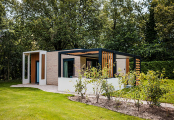 Moderne lodge i naturen med åbent terrasseområde, grønne omgivelser og store træer i baggrunden.