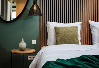 Moderne lodgeværelse med træpaneler, seng med grøn pude, natbord, vase, spejl og hængelampe.