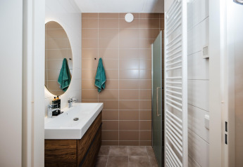 Moderne badeværelse på lodge med brun flisevæg, hvid vask, træskab, spejl og grønne håndklæder.
