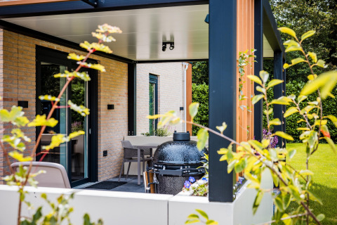 Moderne lodge med overdækket terrasse, udendørs spisebord og grill, omgivet af grøn have og planter.