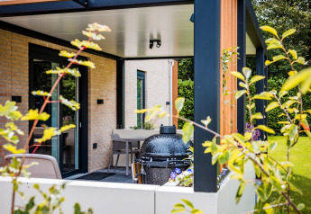 Moderne lodge med overdækket terrasse, udendørs spisebord og grill, omgivet af grøn have og planter.