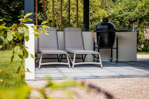 Twee ligzetels en een barbecue op een terras bij een lodge, omringd door een groene tuin.
