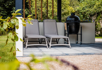 To liggestole og en grill på en terrasse i fredelige omgivelser ved en lodge med grøn havebaggrund.