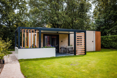 Moderne lodge met overdekte terras, zithoek buiten, gazon en omringende bomen in het daglicht.