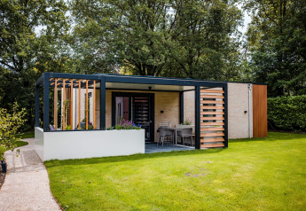 Moderne lodge med overdækket terrasse, omgivet af grøn græsplæne og træer, set i dagslys.