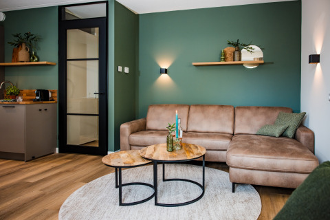 Salotto moderno da lodge con parete verde, divano angolare beige, tavolini rotondi e mensola decorata.