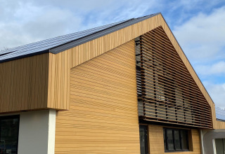 Moderne houten lodge met hellende lamellen en donker dak onder een deels bewolkte hemel.