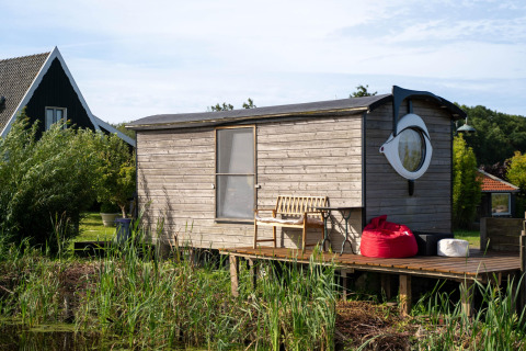 Kleines Holzhaus mit runden Fenstern und Terrasse am Wasser, umgeben von Pflanzen in den Niederlanden.
