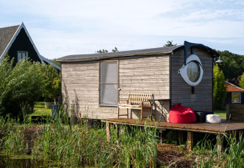 Pequeña casa de madera con ventana redonda y terraza junto al agua, rodeada de plantas en Países Bajos.