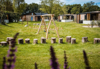 Grøn legeplads med trægynger og stubber foran moderne lodges i naturskønne omgivelser.