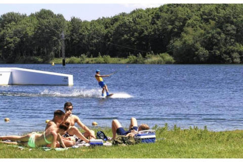 Mensen relaxen aan het gras bij een meer, terwijl iemand wakeboardt in de verte bij de bomen.