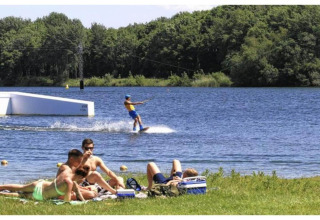 Persone che si rilassano sull'erba vicino a un lago, con una persona che fa wakeboard sullo sfondo.